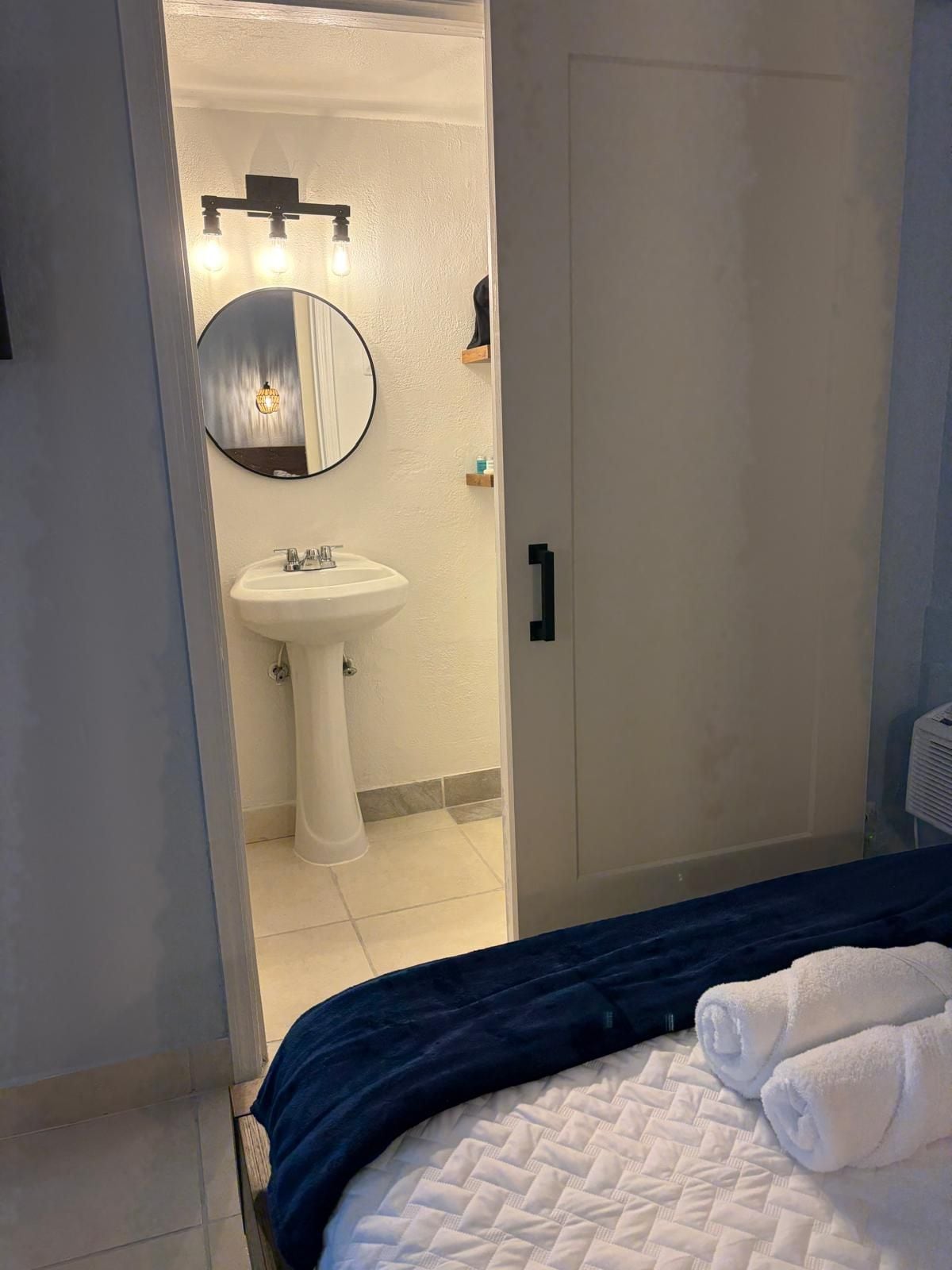 A bedroom with a bed, a bedside table, and a view into a bathroom with a sink and mirror.