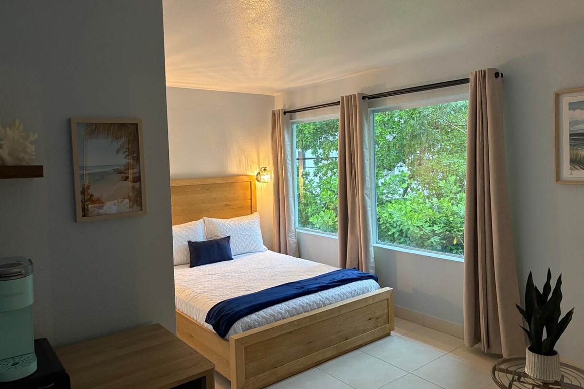 Un dormitorio con cama, mesita de noche, ventana con cortinas y vistas al exterior.