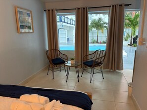 A hotel room with a bed, two wicker chairs, a small table with a plant, and a view of a swimming pool and palm trees. - Beautiful Deluxe Queen Room (Fort Lauderdale)