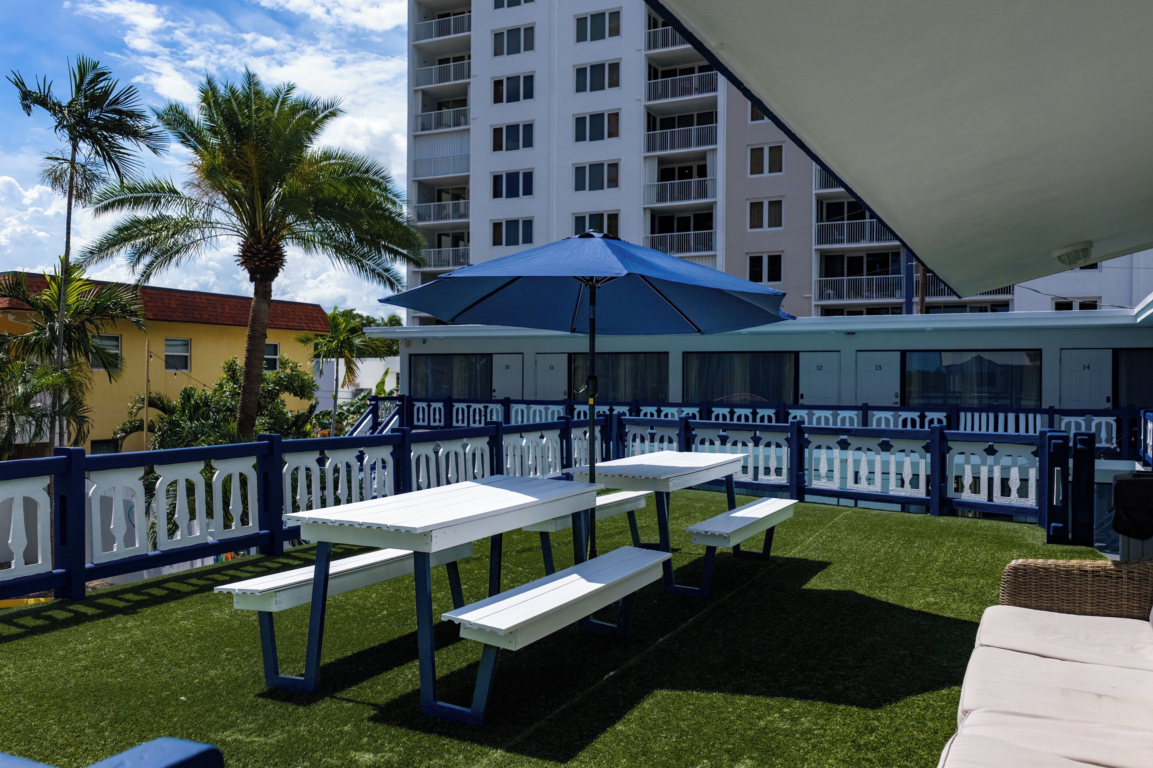Un espace extérieur aménagé en terrasse, avec des tables et des bancs blancs, un parasol bleu et une clôture blanche.