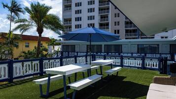 Un espace extérieur aménagé en terrasse, avec des tables et des bancs blancs, un parasol bleu et une clôture blanche.