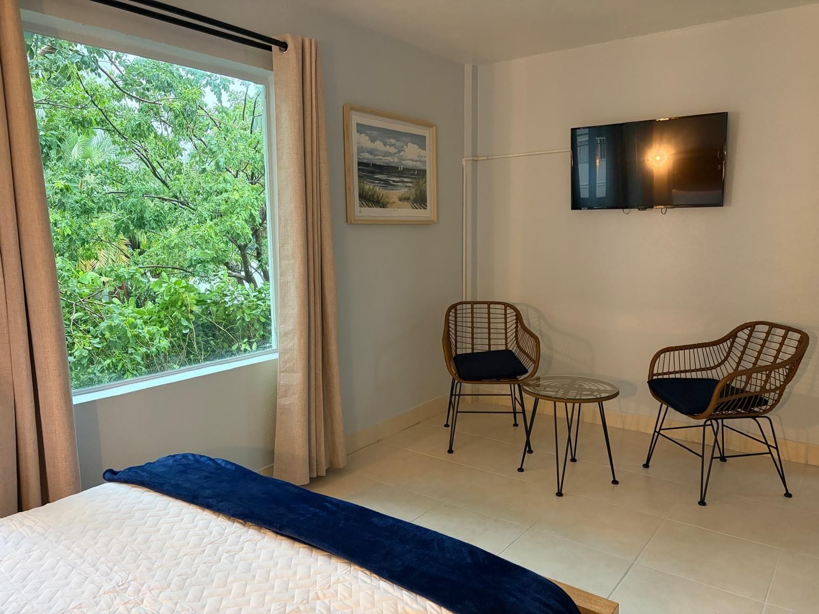 Una habitación con una cama, dos sillas de mimbre, una mesita, un televisor y un ventanal con cortinas.