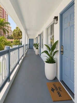 Corredor coberto com porta azul, plantas em vasos e tapete de entrada.