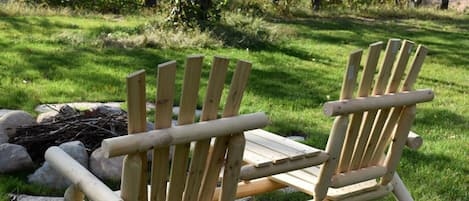 Two wooden Adirondack chairs in a grassy outdoor area with a fire pit in the background.