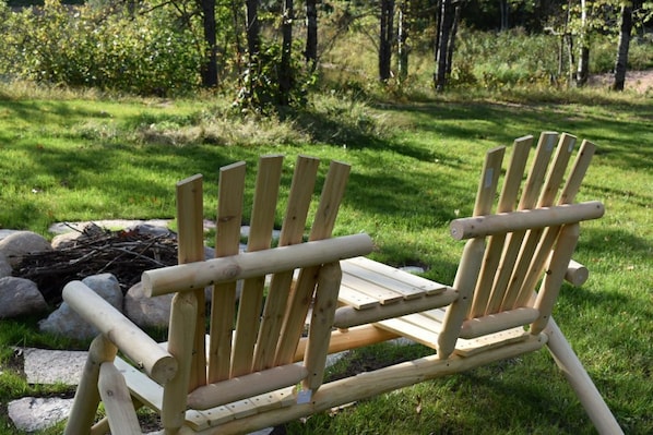 Two wooden Adirondack chairs in a grassy outdoor area with a fire pit in the background.