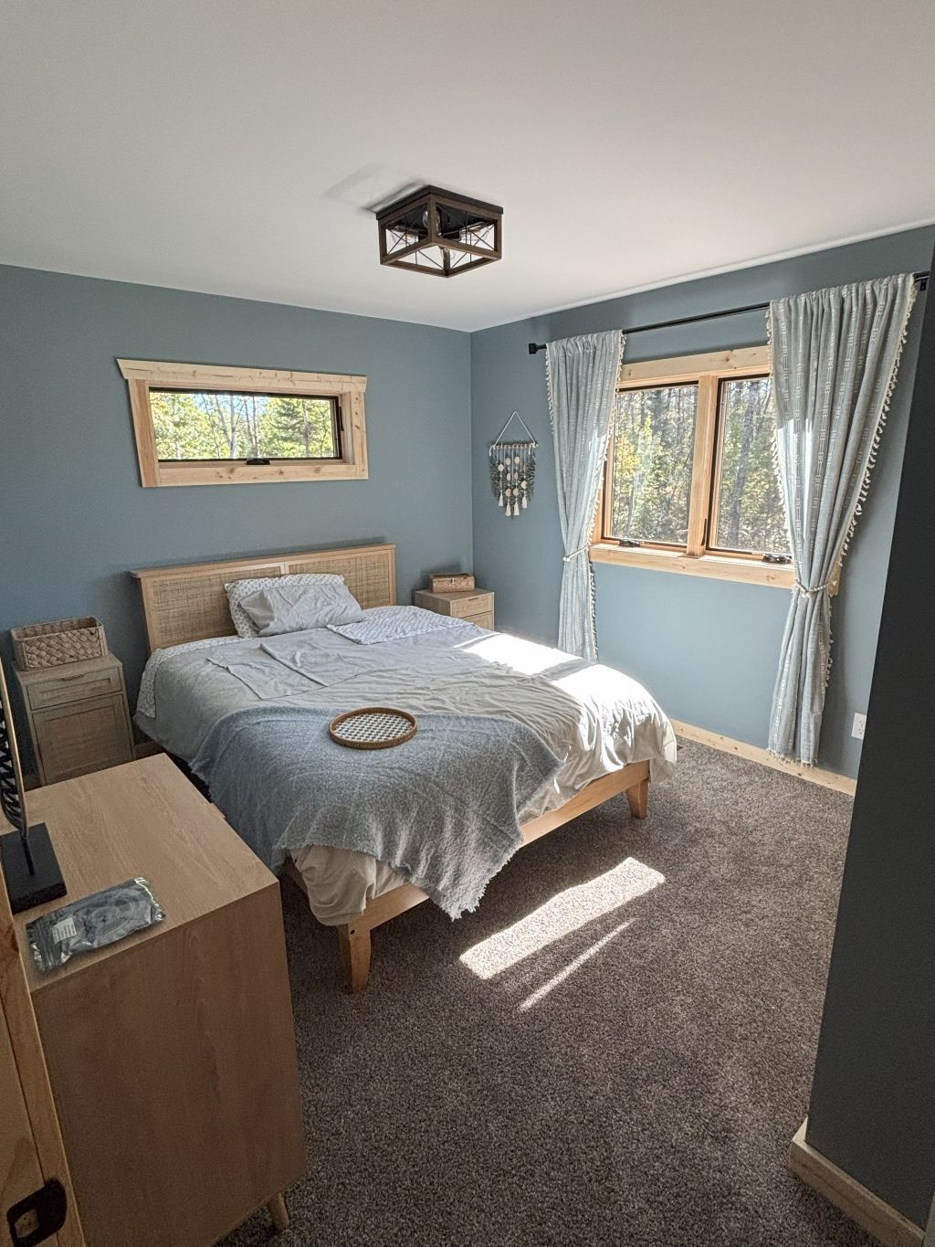 A bedroom with a bed, wooden headboard, bedside tables, a window with curtains, and a wall-mounted light fixture.