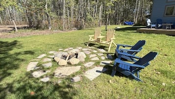 A backyard with a stone fire pit, two Adirondack chairs, and a deck with outdoor furniture.