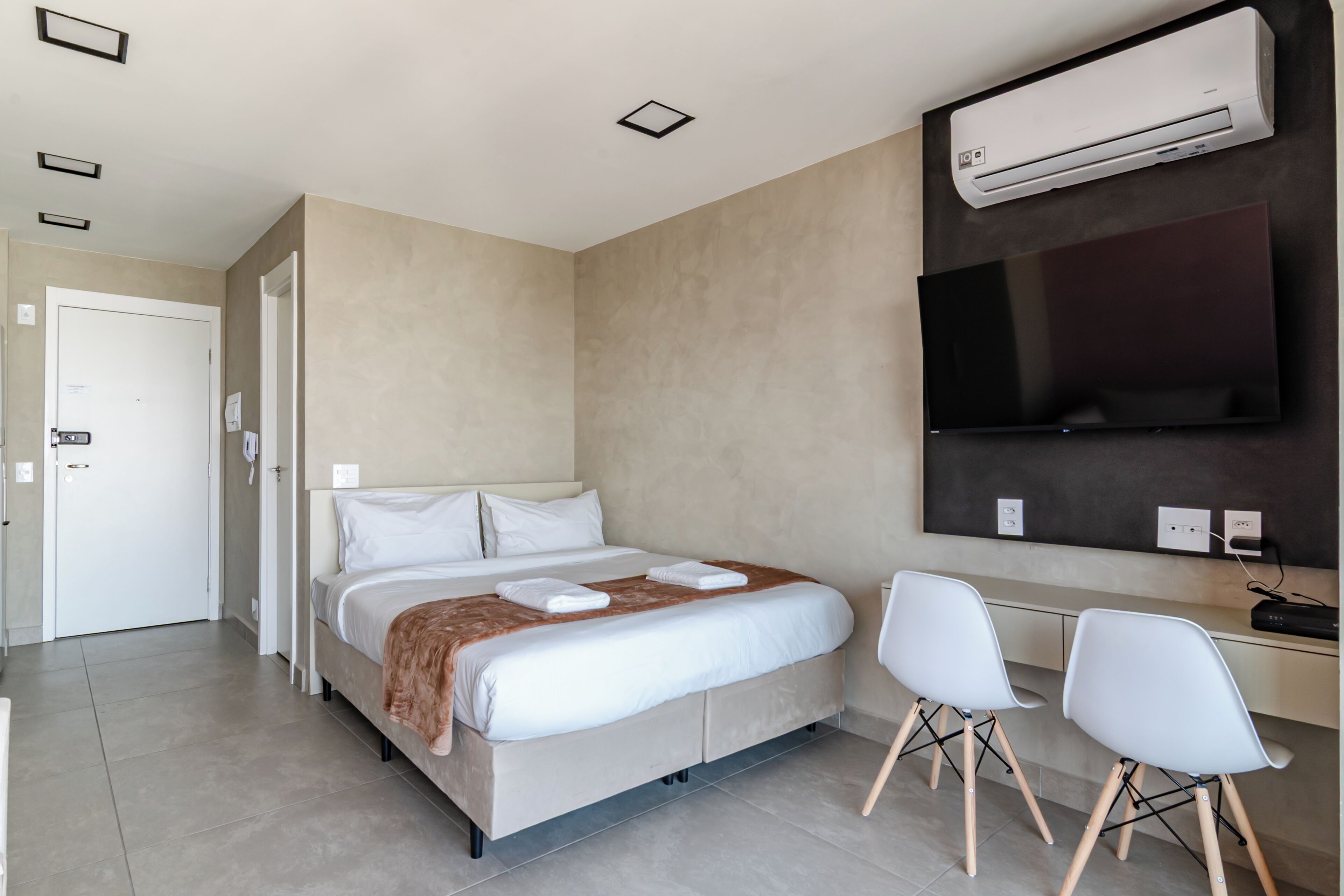 Quarto de hotel com cama, escrivaninha com cadeiras, televisão de tela plana e ar condicionado.