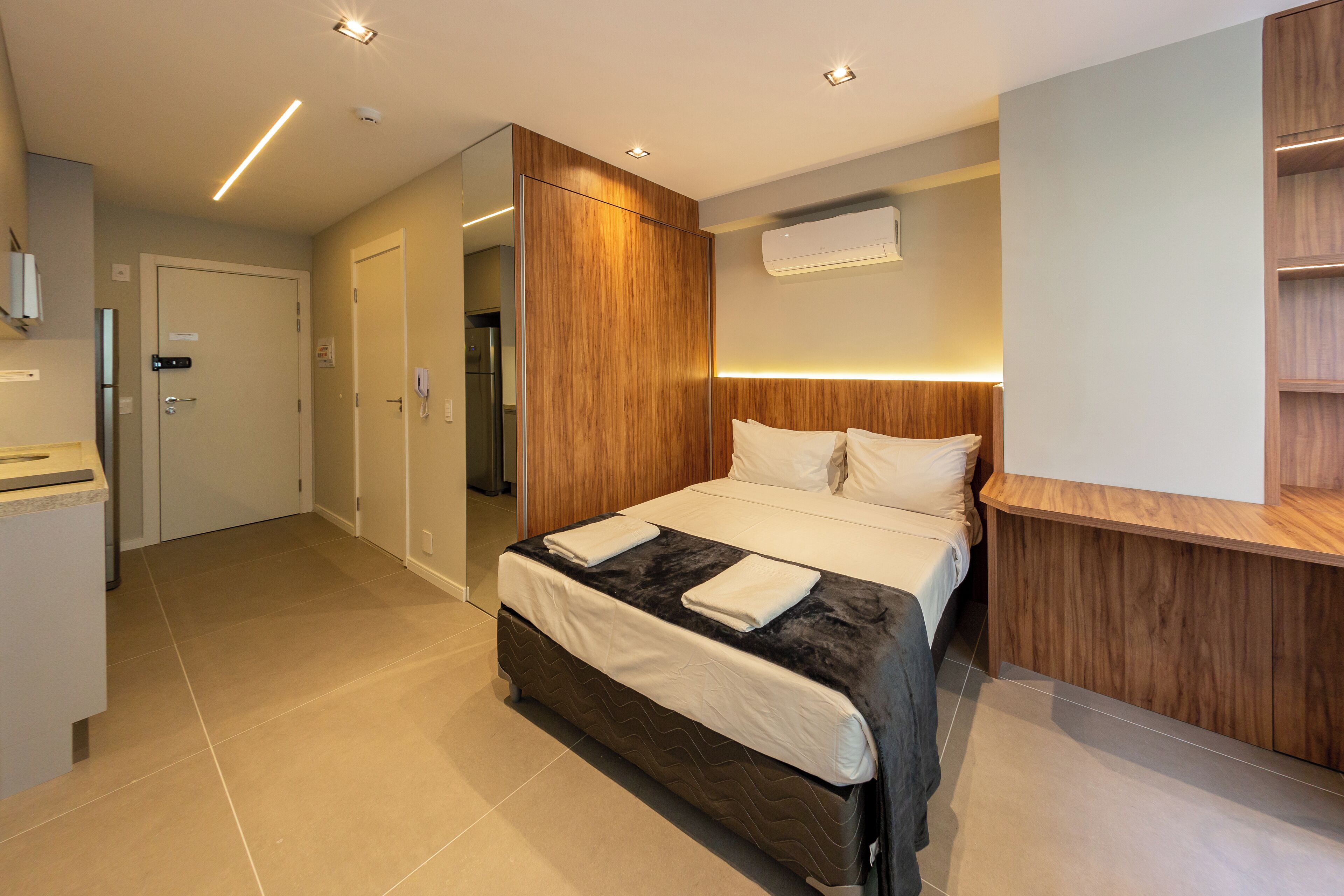 Quarto de hotel moderno com cama, cabeceira de madeira, prateleira embutida e banheiro visível por uma porta aberta.