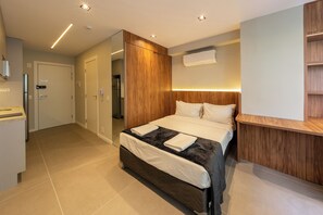 Quarto de hotel moderno com cama, cabeceira de madeira, prateleira embutida e banheiro visível por uma porta aberta.