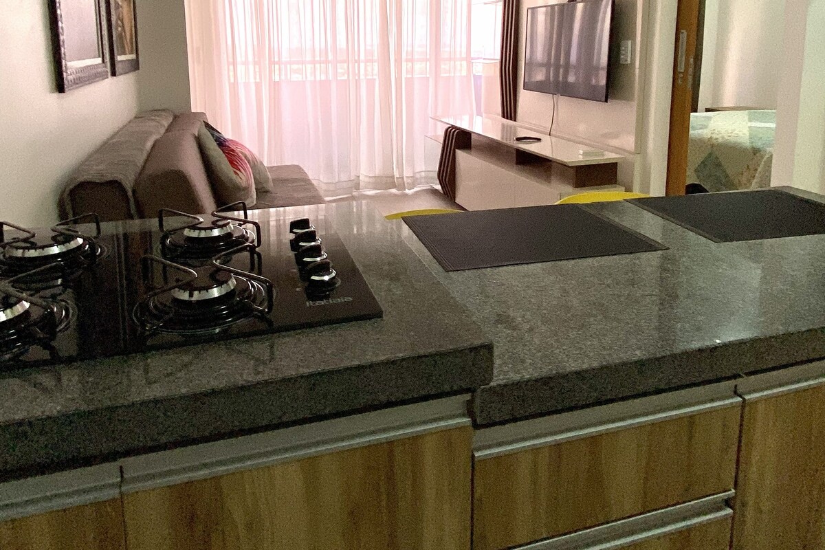 A modern kitchen with a gas stove, a dining area with a sofa, and a TV mounted on the wall.