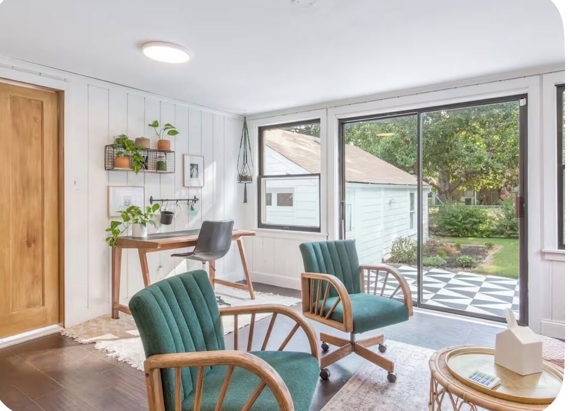 A room with a wooden door, a desk with a chair, two green upholstered chairs, a wicker side table, and a view of a garden through a glass door.