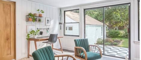A room with a wooden door, a desk with a chair, two green upholstered chairs, a wicker side table, and a view of a garden through a glass door.