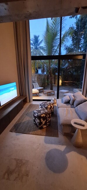 A modern living room with a large TV, comfortable seating, and a view of palm trees. - Komorebi House - Episode 6 (Cabarete)