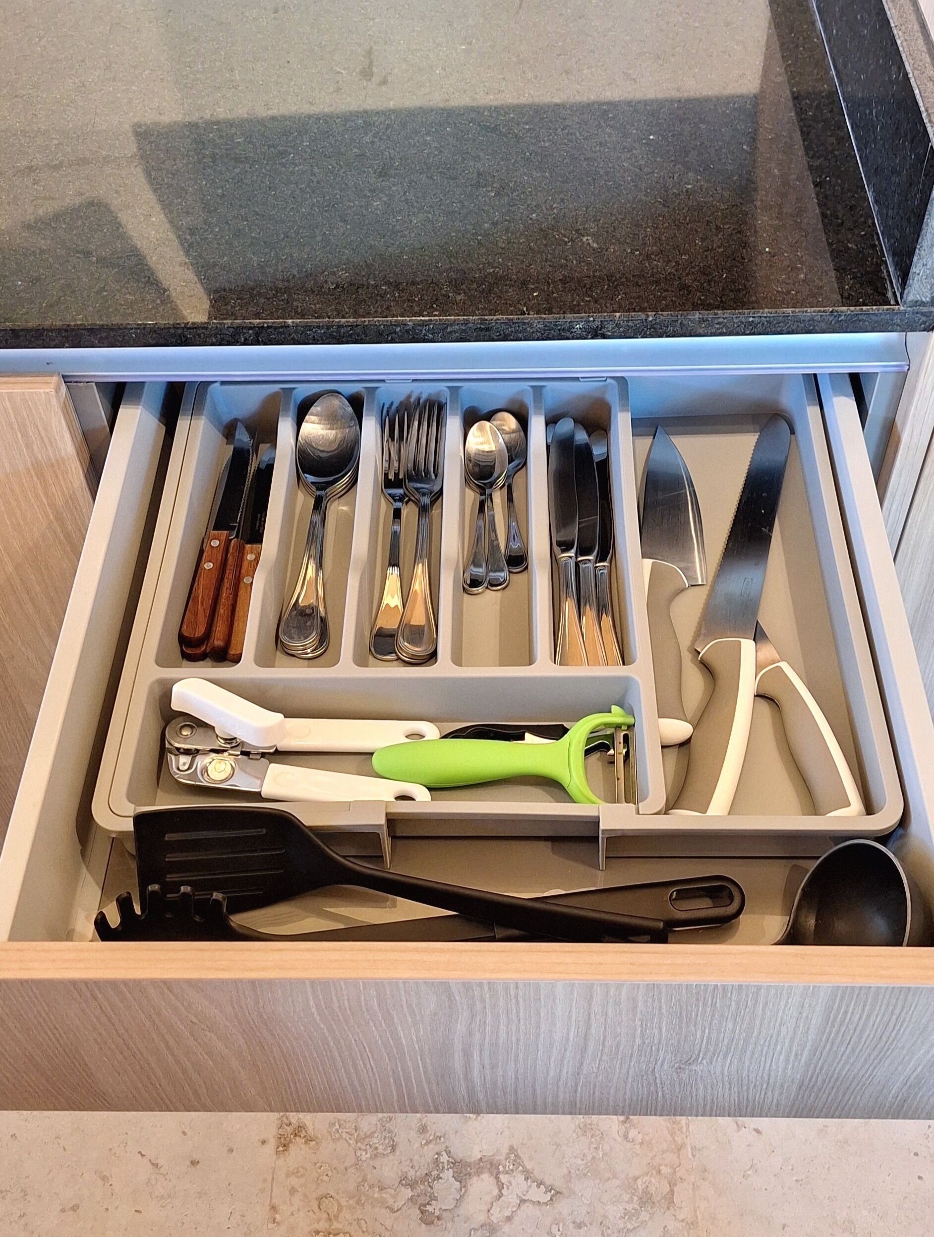 A drawer with kitchen utensils including spoons, forks, knives, and a green-handled peeler.