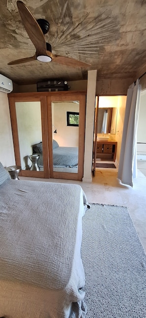 Quarto com cama, ventoinha no teto, banheiro com pia e espelho, e vista para o exterior.