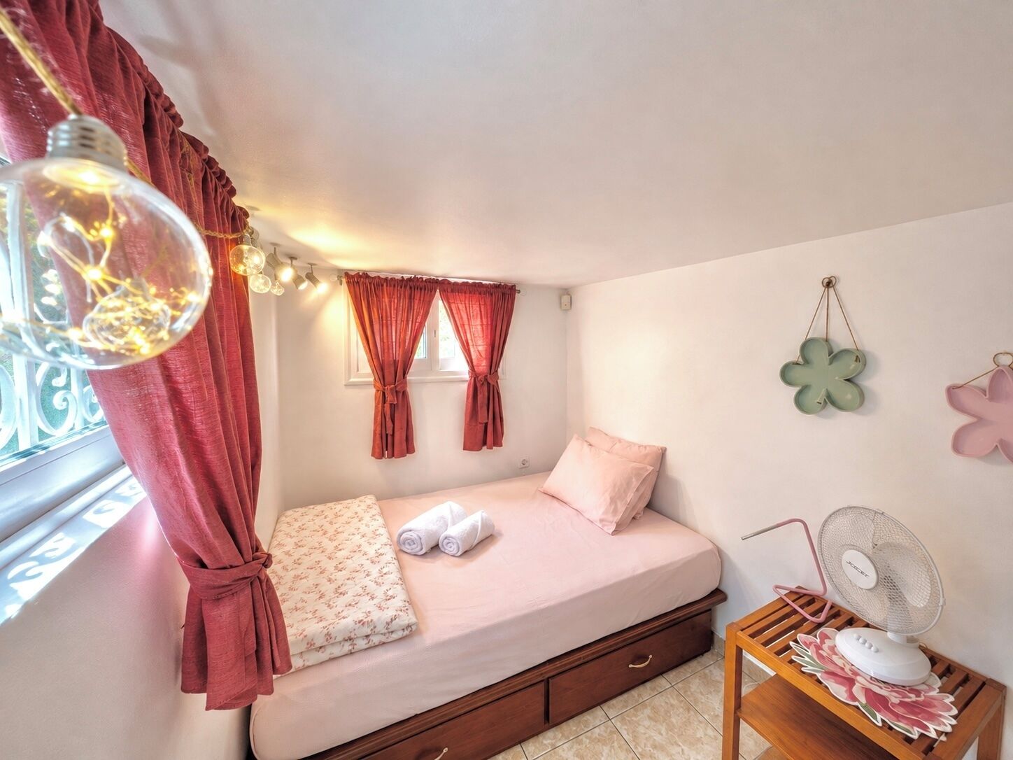 A bedroom with a bed, a window with curtains, a bedside table with a fan, and wall decorations.