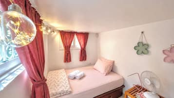 A bedroom with a bed, a window with curtains, a bedside table with a fan, and wall decorations.