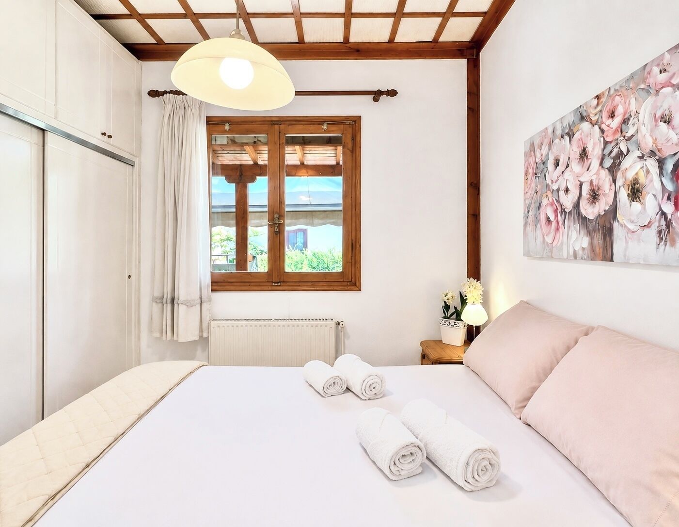 A bedroom with a bed, a window with curtains, a radiator, a nightstand with a lamp, and a floral painting on the wall.