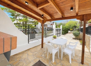 A covered patio with a stone floor, white plastic chairs, a table, and a grill.