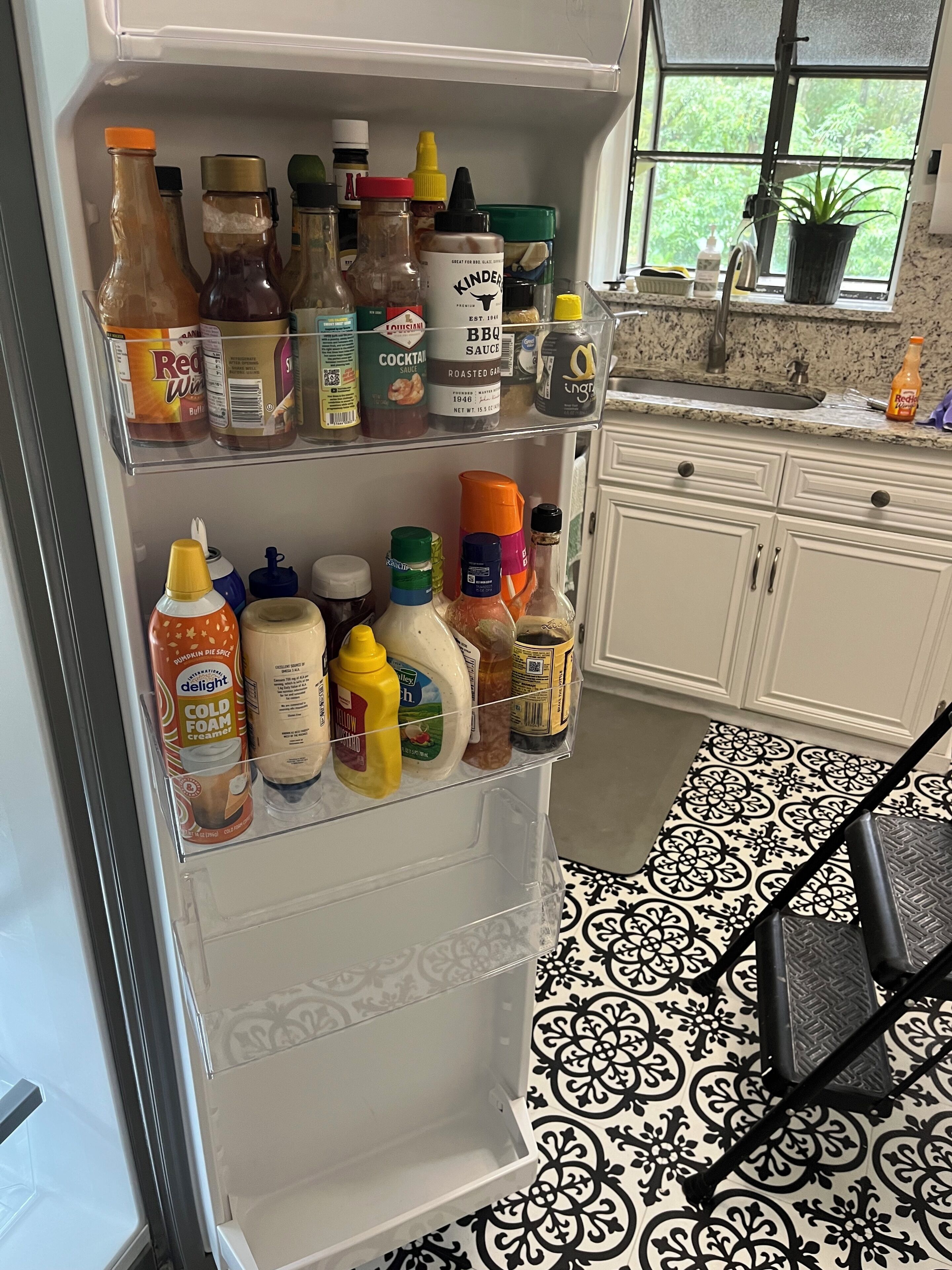 A kitchen with a refrigerator stocked with condiments and a window with a view of greenery.