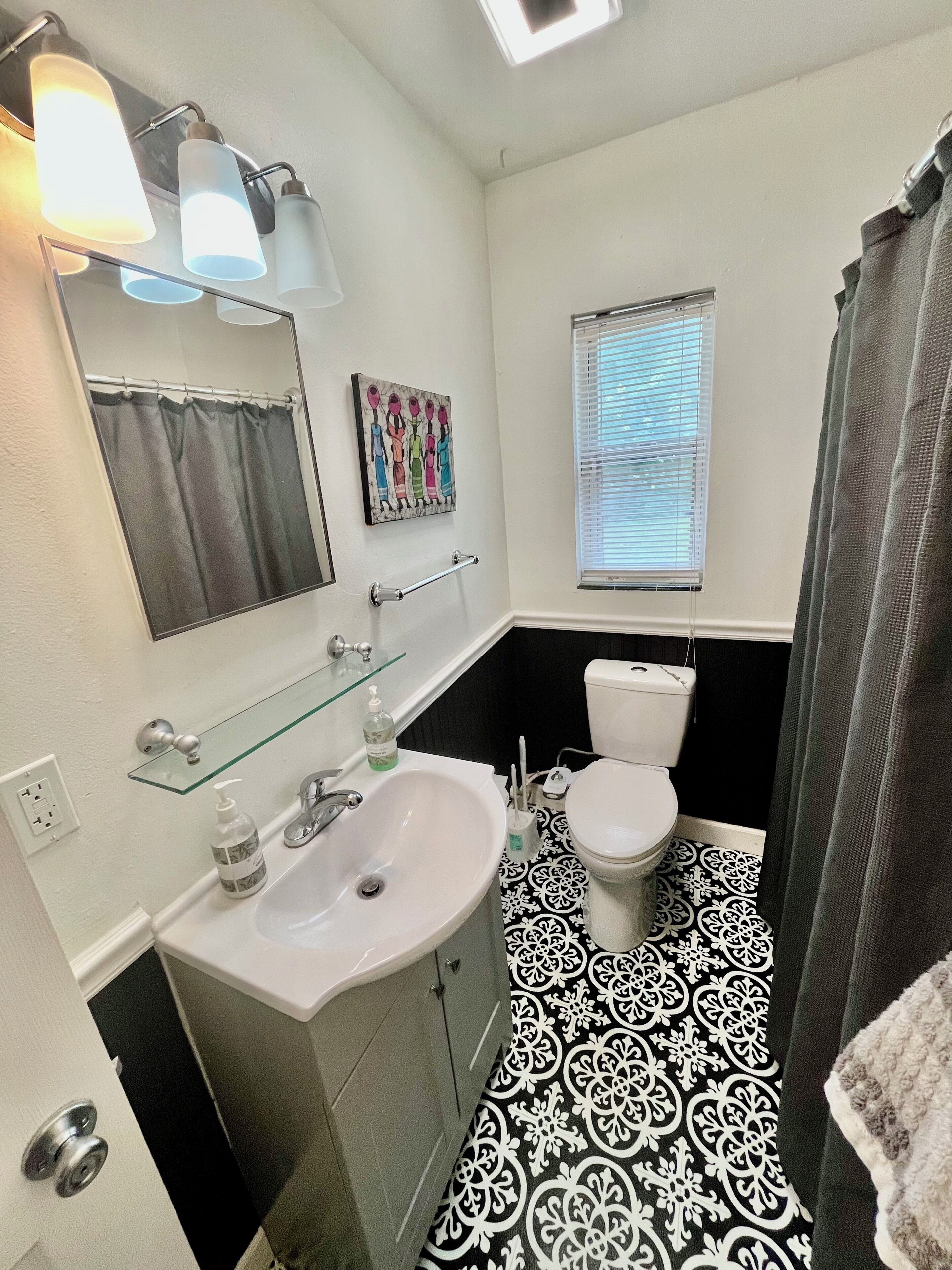 A bathroom with a white sink, a toilet, a glass shelf with toiletries, a mirror, and a window with blinds.