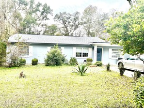 Uma casa térrea com fachada azul, porta da frente e pequeno jardim frontal com grama e alguns arbustos.