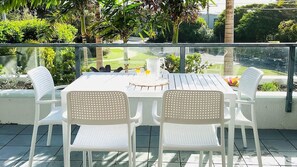 Une terrasse extérieure avec une table et quatre chaises, entourée d’une rambarde en verre, donnant sur un jardin avec différentes plantes et offrant une vue sur la rue.