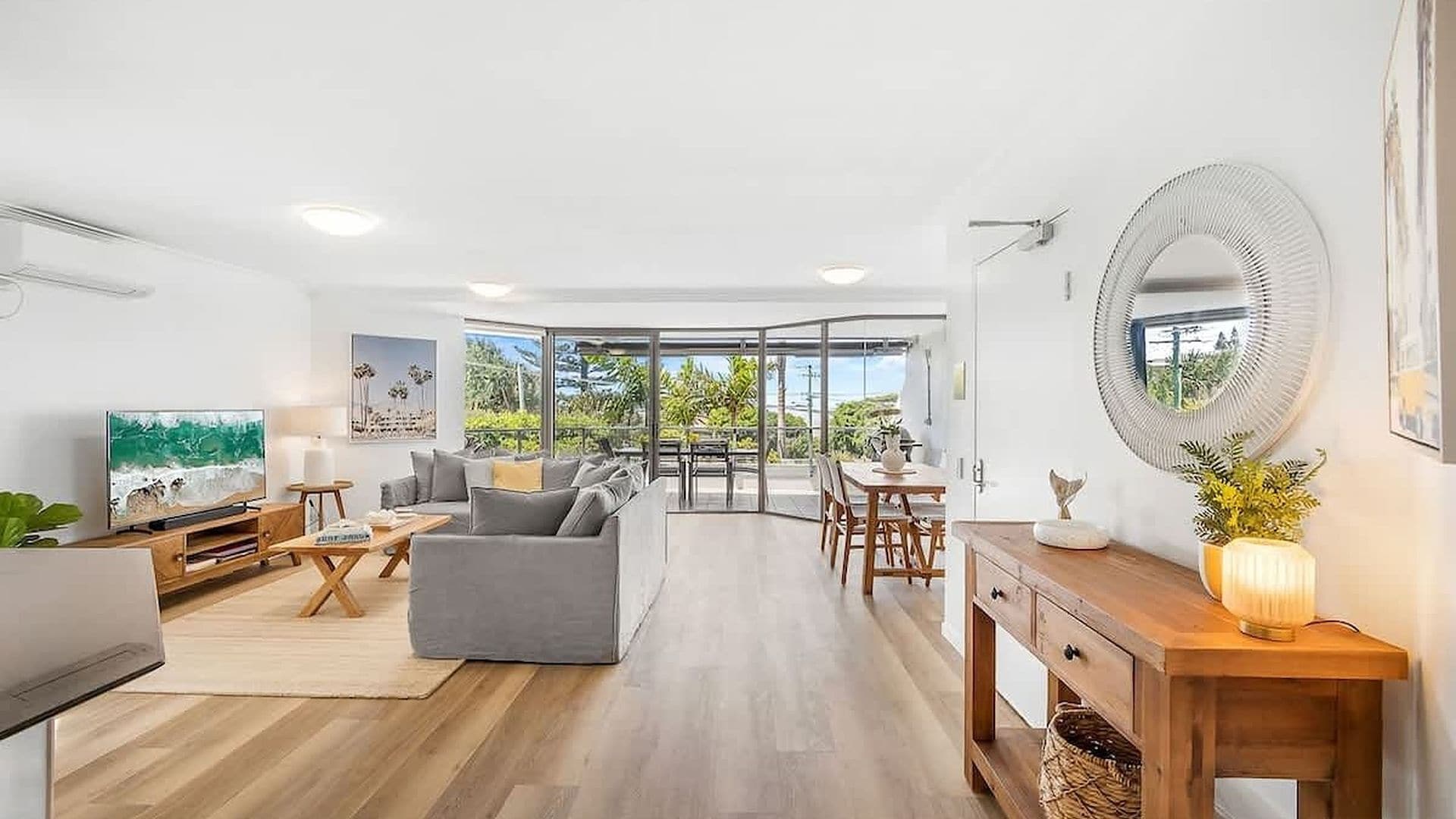 A modern living room with a grey sofa, wooden coffee table, and a TV on the left. A dining area with a wooden table and chairs is in the center. The room has large windows and a view of greenery outside.