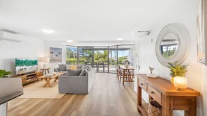 A modern living room with a grey sofa, wooden coffee table, and a TV on the left. A dining area with a wooden table and chairs is in the center. The room has large windows and a view of greenery outside. - Belmore Palms Sunshine Beach (Sunshine Beach)
