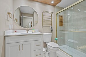 Une salle de bain moderne avec un meuble-lavabo blanc, un miroir rond, des toilettes et une douche séparée par une paroi de verre.