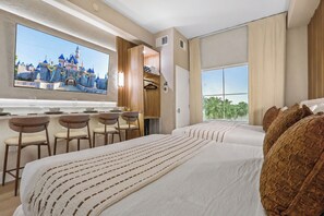 Une chambre d’hôtel avec un grand lit, un bureau avec des chaises, un téléviseur diffusant une image de château et une fenêtre donnant sur des palmiers.