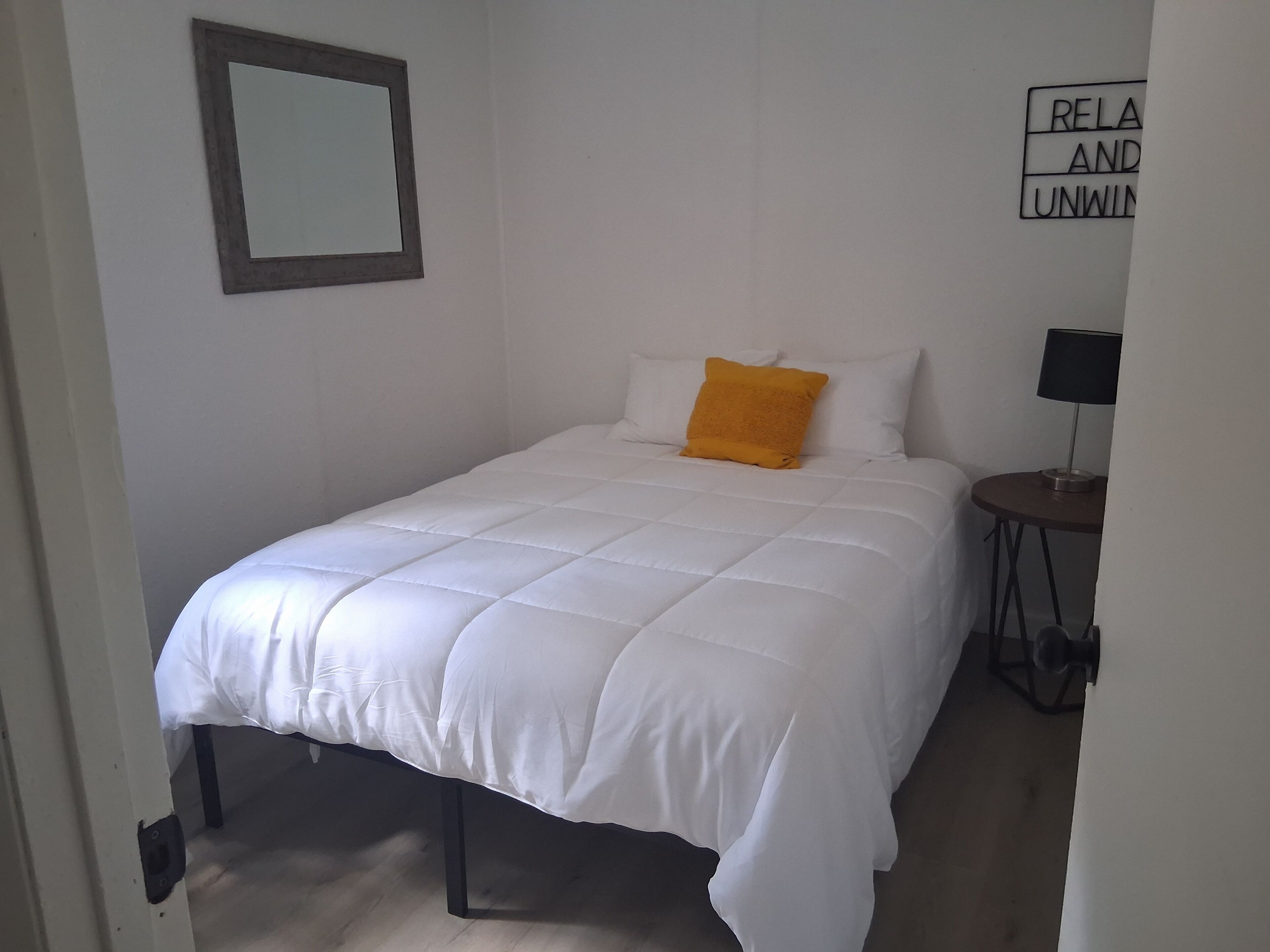 Una cama bien hecha con edredón blanco y una sola almohada naranja, una mesita de noche pequeña con una lámpara, y un letrero en la pared que dice 'RELAX AND UNWIND'.