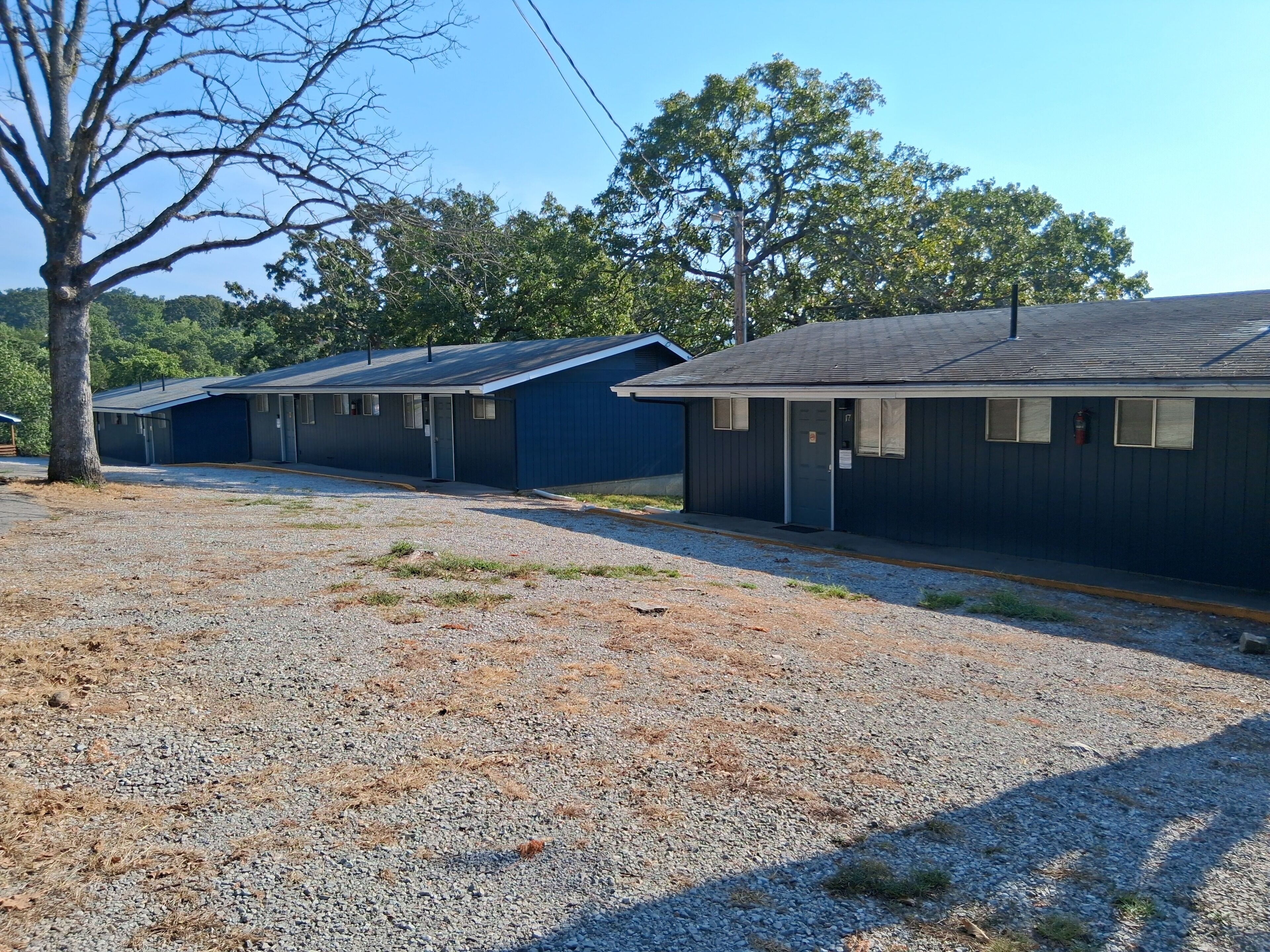 A row of single-story buildings with a gravel driveway and trees in the background.