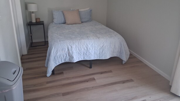 A bedroom with a bed, a nightstand, a lamp, and a trash bin.