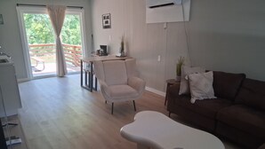 A living room with a sofa, a chair, and a unique white coffee table. There is a balcony with a wooden railing and a view of greenery.