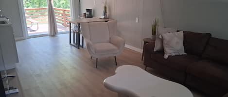 A living room with a sofa, a chair, and a unique white coffee table. There is a balcony with a wooden railing and a view of greenery.