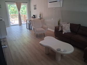 A living room with a sofa, a chair, and a unique white coffee table. There is a balcony with a wooden railing and a view of greenery.