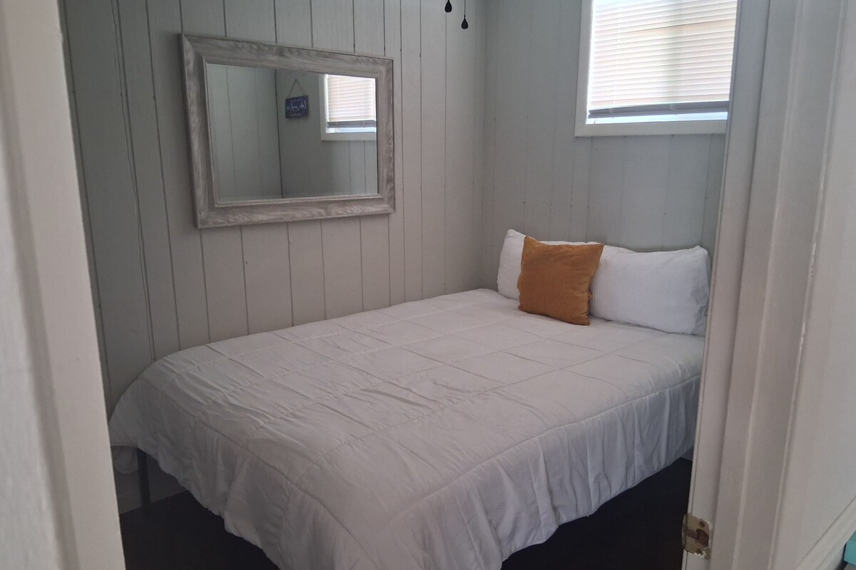 A small, single-bed room with a white bedspread, a brown pillow, a mirror on the wall, and a window with blinds.