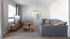En moderne stue med en grå sofa, et rundt sofabord, et fjernsyn på et træstel og et vindue med gardiner.