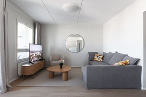 A modern living room with a grey sofa, a round coffee table, a TV on a wooden stand, and a window with curtains. - Stylish 2-bedroom family apartment w/ sauna, parking (Helsinki)