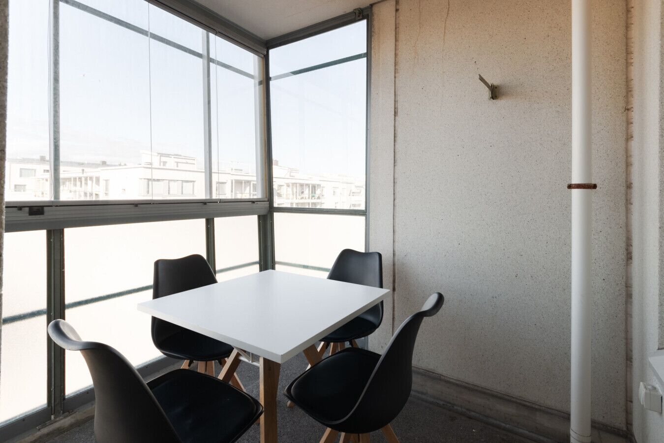 A conference room with a white table and four black chairs, large windows, and a plain wall.
