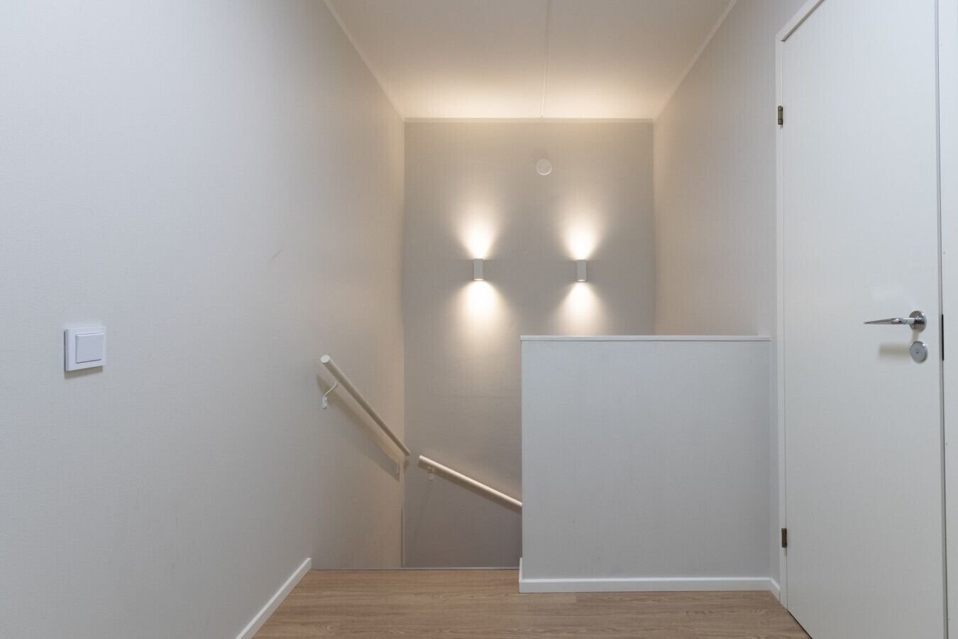 A narrow hallway with a wooden floor, a white door on the right, and a light switch on the left.