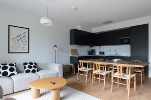 Moderni olohuone, jossa on sohva, sohvapöytä, ruokapöytä ja tuolit. Keittiöalueella on kaappeja ja kodinkoneita.