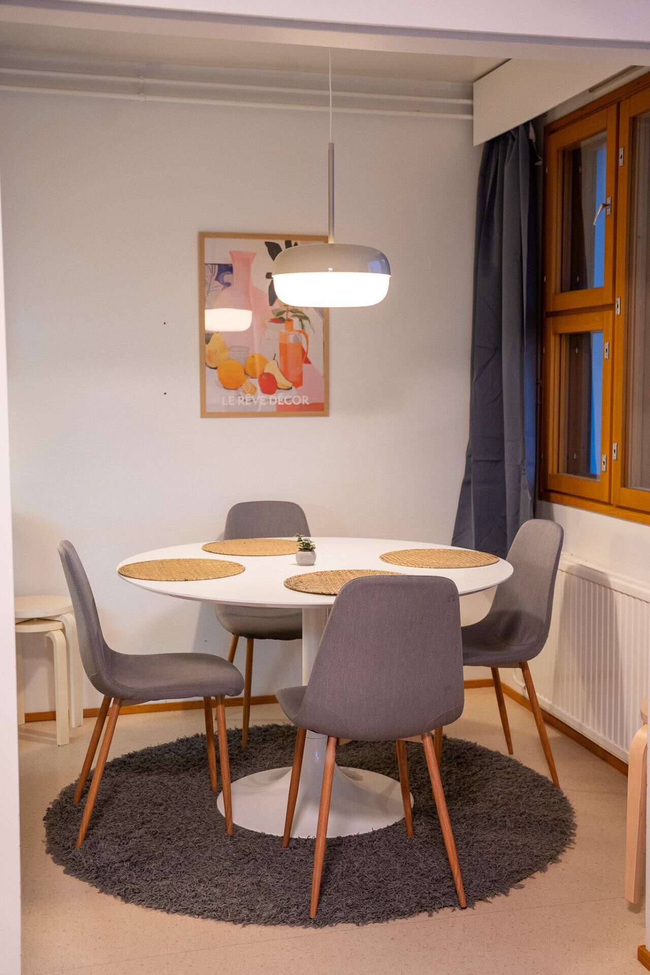 A dining area with a round table, four grey chairs, a small plant, and a hanging light fixture.