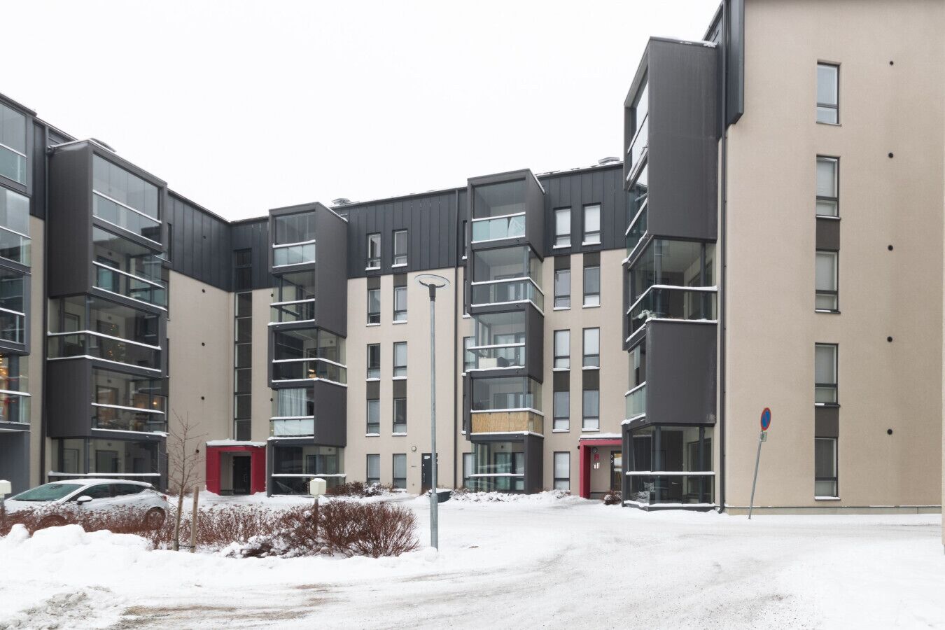 Kerrostalo, jossa on useita kerroksia, parvekkeita, pysäköintialue ja lumipeitteinen maa.
