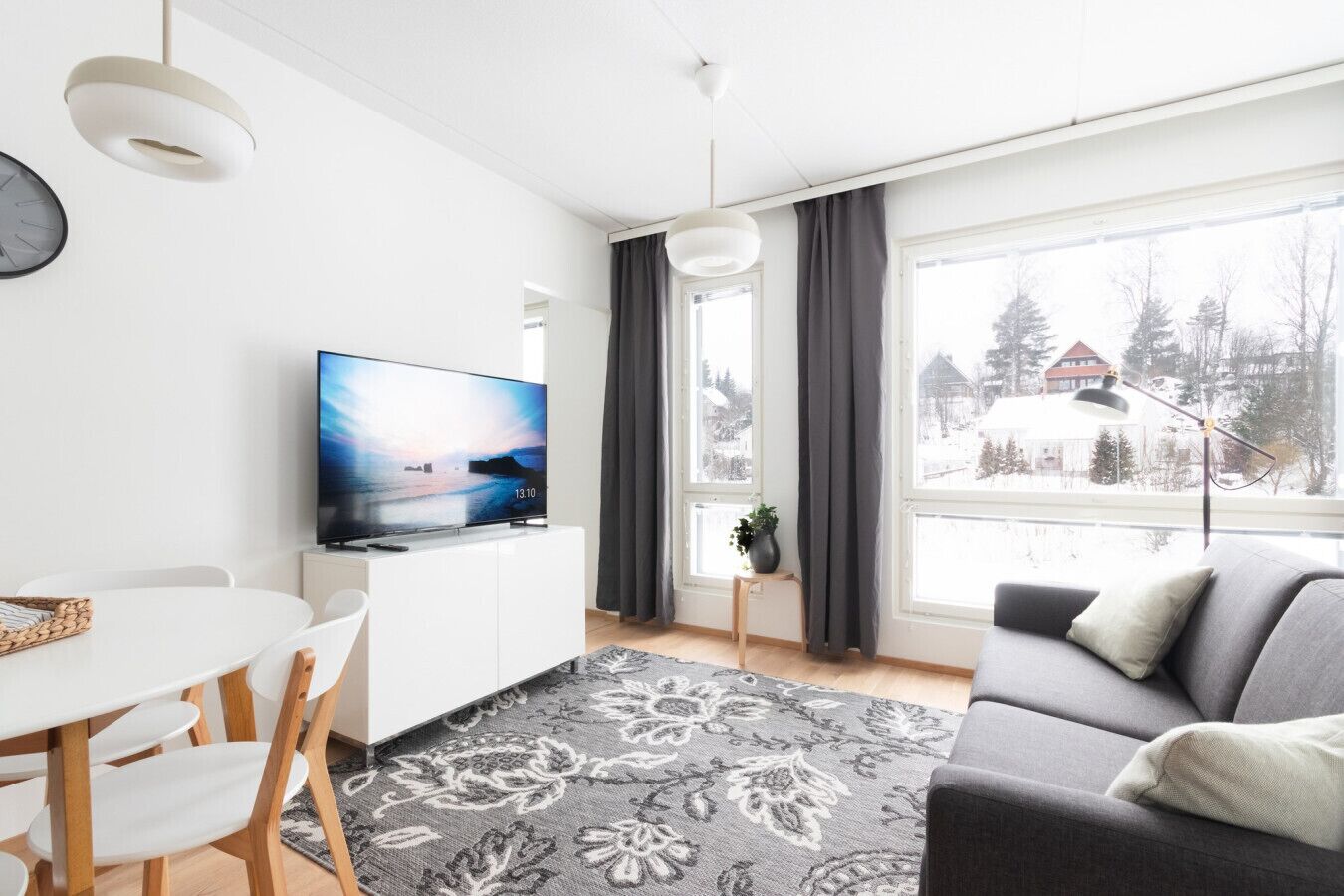 Ein modernes Wohnzimmer mit einem Flachbildfernseher, einem Essbereich mit einem weißen Tisch und Stühlen, einem grauen Sofa und einem großen Fenster mit Blick auf eine verschneite Landschaft.