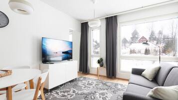 Ein modernes Wohnzimmer mit einem Flachbildfernseher, einem Essbereich mit einem weißen Tisch und Stühlen, einem grauen Sofa und einem großen Fenster mit Blick auf eine verschneite Landschaft.