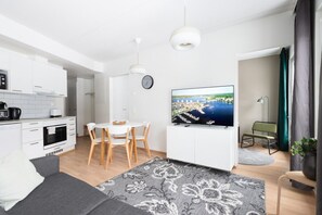 Moderni olohuone, jossa on litteänäyttöinen televisio, ruokailutila pöytineen ja tuoleineen, sohva sekä keittiö valkoisilla kaapeilla ja kodinkoneilla.