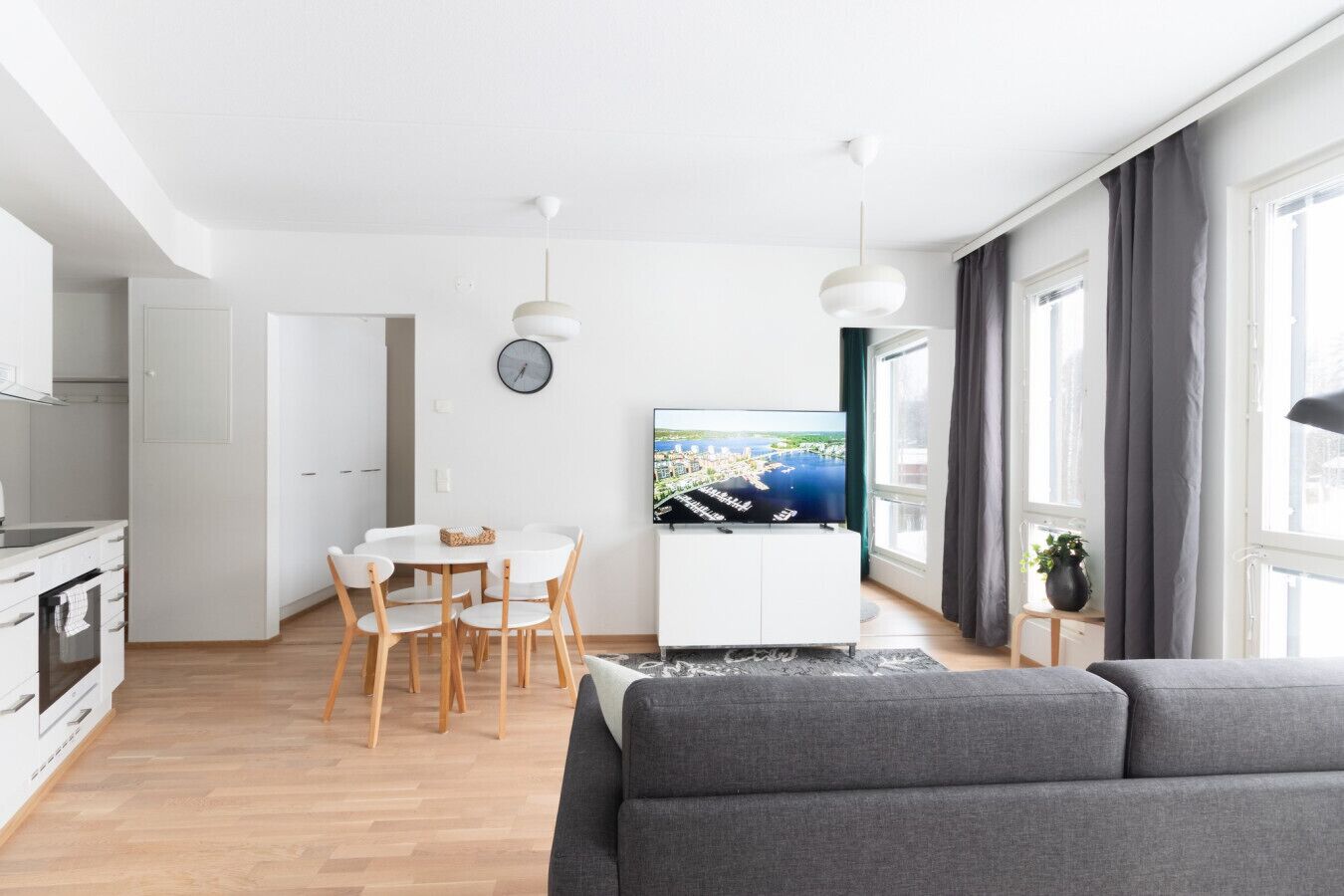 Moderni olohuone, jossa on harmaa sohva, valkoisella pöydällä ja tuoleilla varustettu ruokailutila, valkoisilla kaapeilla varustettu keittiö ja televisio, jossa näkyy kaupunkimaisema.